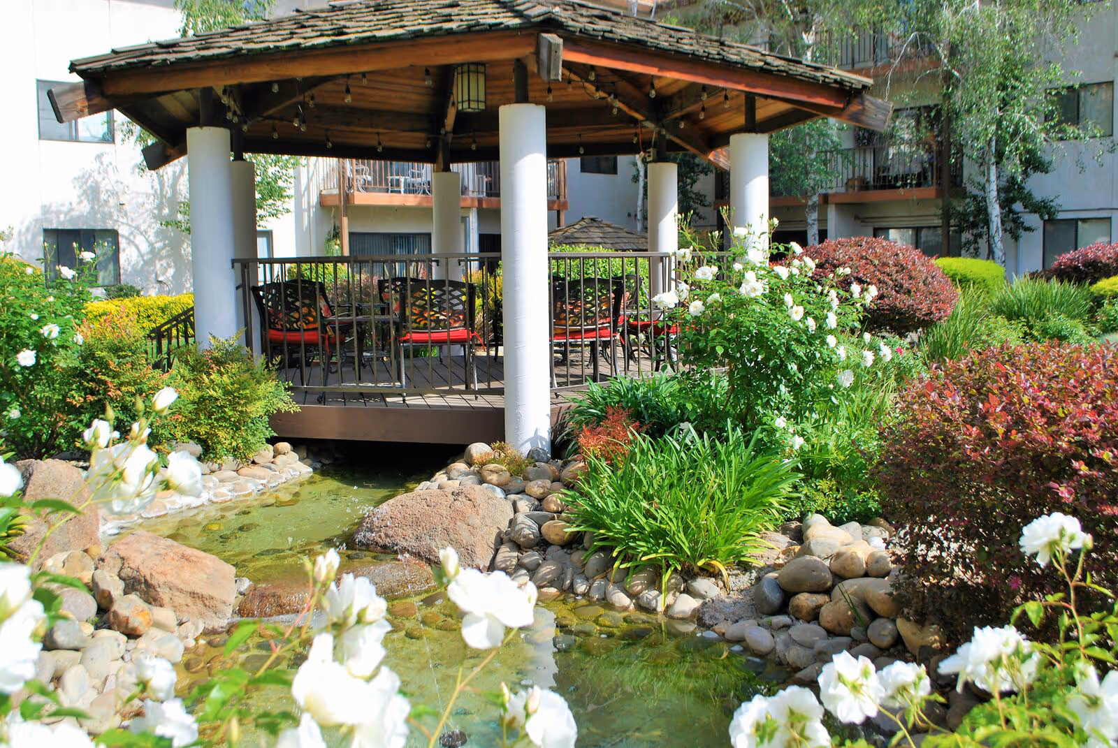A gazebo with a wooden roof and white pillars surrounded by lush greenery and blooming white flowers. There is a small stream with rocks flowing beneath the gazebo. In the background, parts of a senior living facility building with balconies are visible.