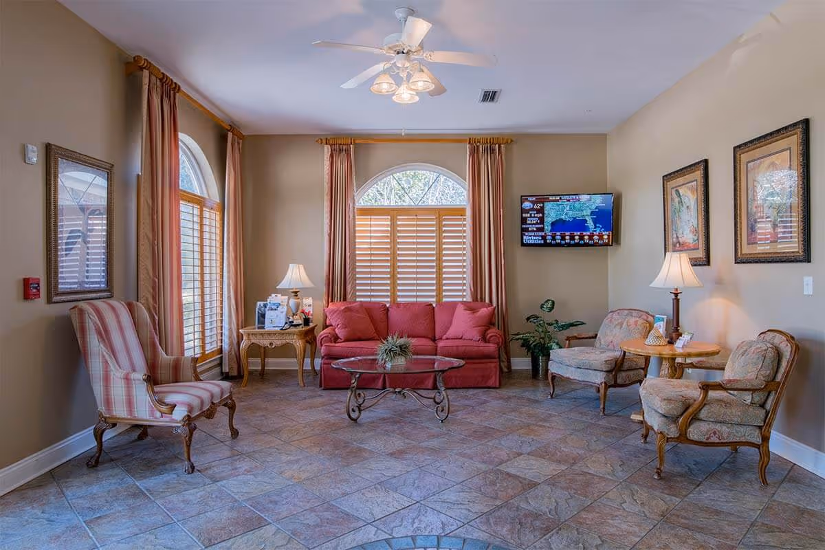 Bright senior living common room with a red sofa, several upholstered chairs, coffee table, side tables, lamps and a wall-mounted TV.