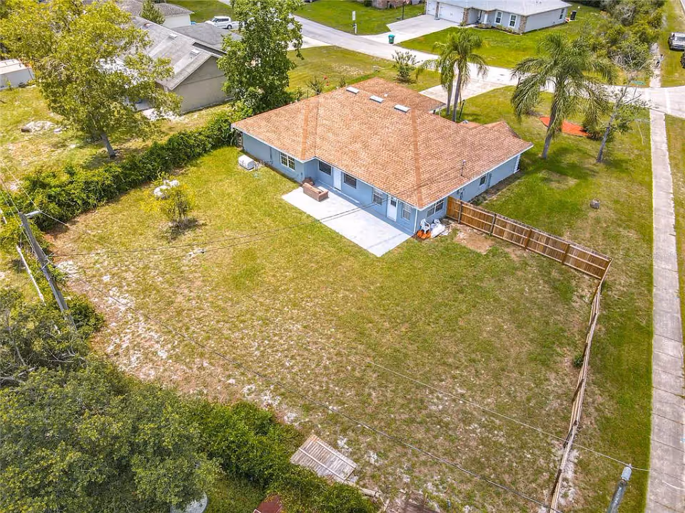 Aerial view of a single-story house with a brown shingle roof and light blue exterior walls, surrounded by a large grassy yard with some trees and a wooden fence on one side. The house is located in a residential neighborhood with other houses and paved sidewalks visible.
