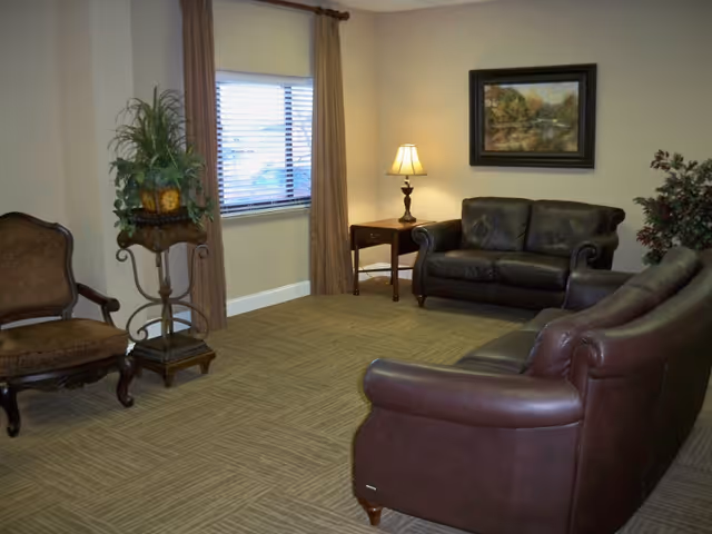 A cozy living room with two dark brown leather sofas, a wooden side table with a lamp, a framed landscape painting on the wall, a window with brown curtains, a decorative plant on a stand, and an upholstered armchair.