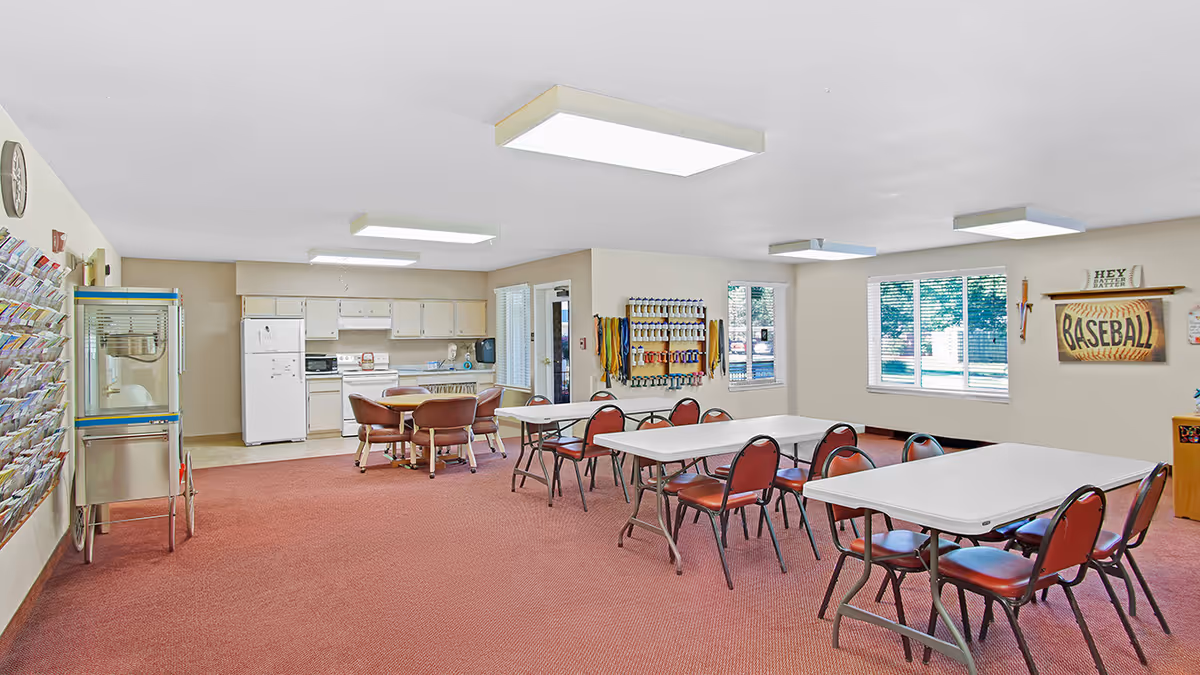 A spacious activity room with red carpet, white folding tables, and red cushioned chairs. The room features a kitchenette with white cabinets, a refrigerator, stove, and microwave. On the left wall, there is a popcorn machine and a rack filled with brochures or pamphlets. The right wall has windows with blinds, a baseball-themed decoration, and a rack holding various exercise bands and equipment. The ceiling has rectangular fluorescent lights.