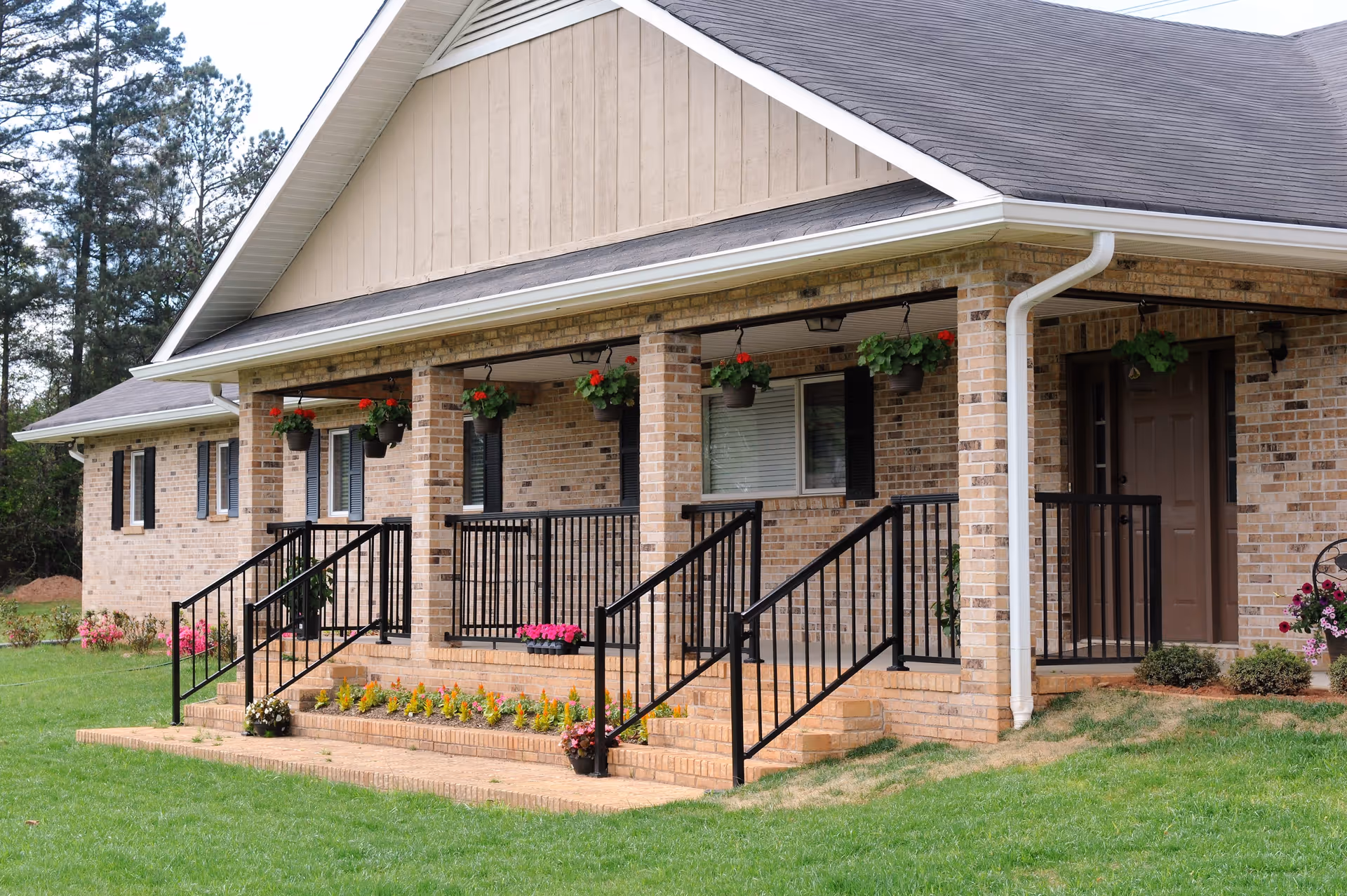 Front exterior view of a single-story brick building with a covered porch featuring black railings and hanging flower pots. There are steps leading up to the porch, and the lawn is well-maintained with flower beds along the front.