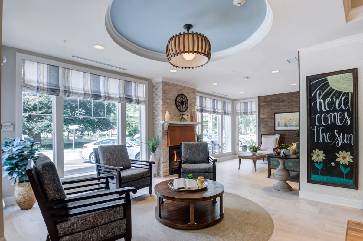 Sunlit seating area and lobby with armchairs around a round coffee table, a fireplace, large windows, and a chalkboard reading "here comes the sun."