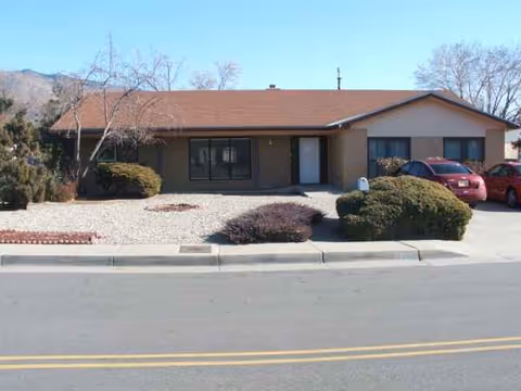 Single-story building with brown exterior, front entrance, and desert landscaping; cars parked on the right.