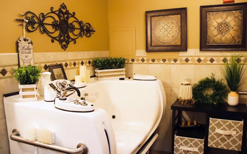 A well-decorated bathroom featuring a white walk-in bathtub with silver handles and controls. The walls are tiled with a decorative border and adorned with framed artwork and a wrought iron wall decoration. There are several green plants and candles placed on and around the bathtub, creating a relaxing atmosphere. A small sign hanging on the wall reads 'Relax & soak'.