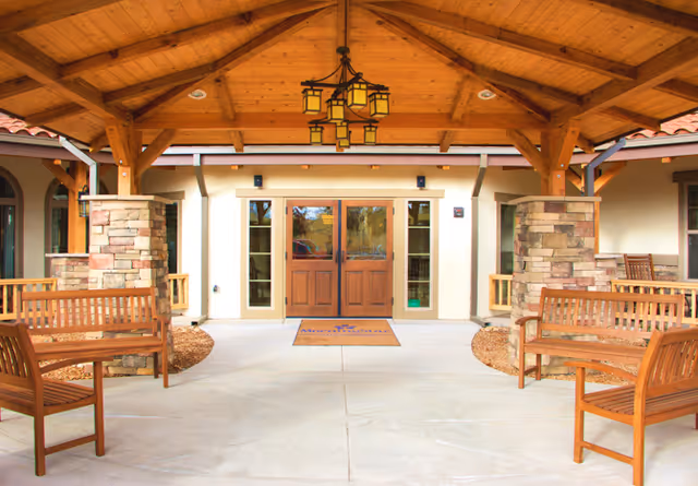 Covered outdoor seating area with wooden benches and stone pillars, leading to double wooden doors of a building under a wooden roof structure with a hanging lantern light fixture.