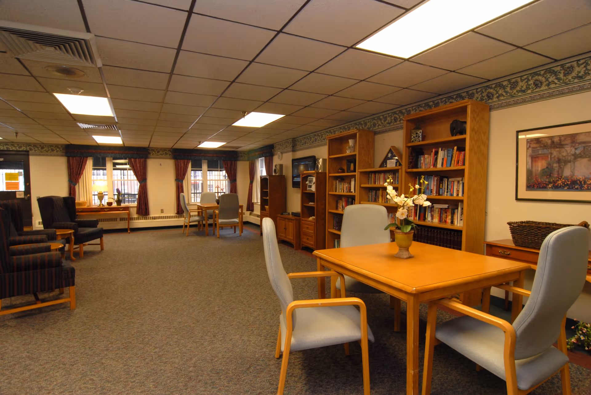 Communal lounge with tables and chairs, bookshelves, and seating in a senior living facility.