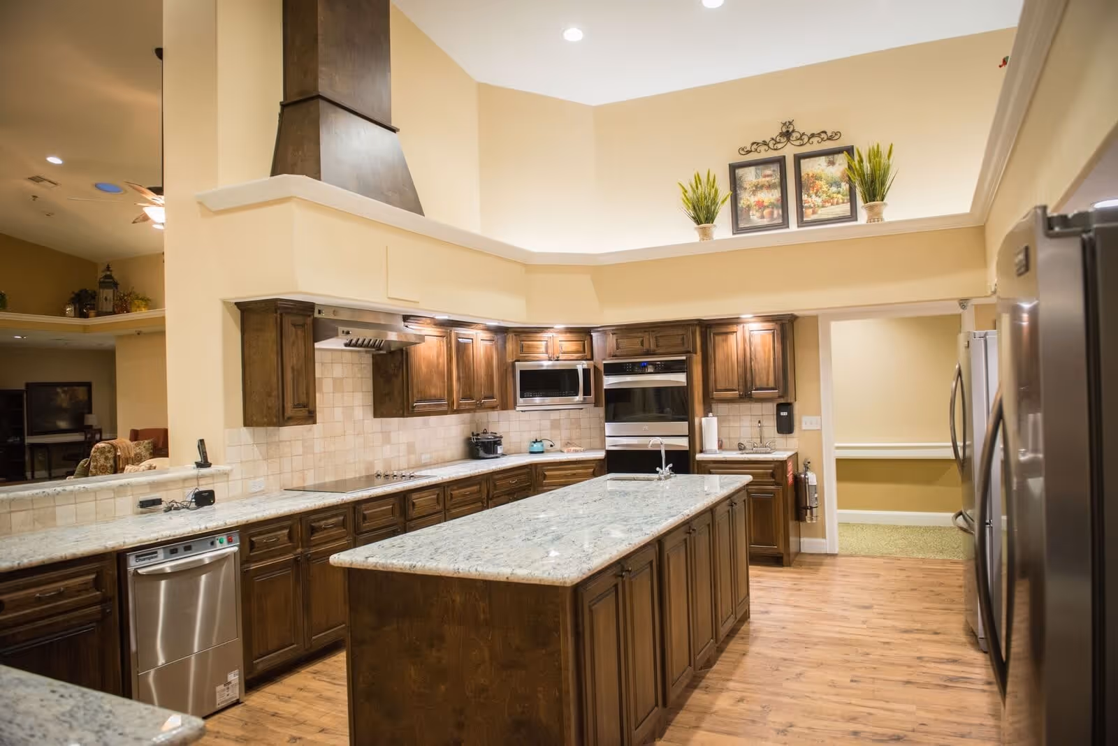 Spacious kitchen with wooden cabinets, a large central island with a granite countertop, stainless steel appliances including a dishwasher, microwave, oven, and refrigerator. The kitchen has a high ceiling with recessed lighting, decorative plants, and framed artwork above the cabinets. The floor is wooden and there is an open doorway leading to another room.
