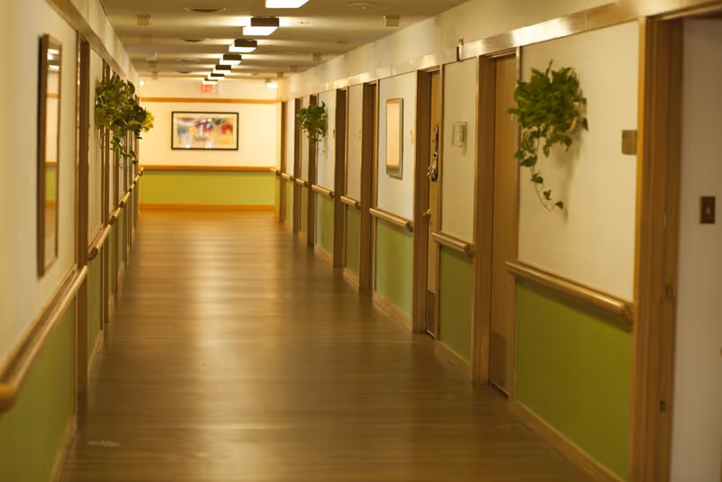 A long, well-lit hallway in a senior living facility with wooden floors and green lower wall panels. Doors line both sides of the hallway, each with a small plant hanging beside it. A framed picture is visible at the end of the corridor.