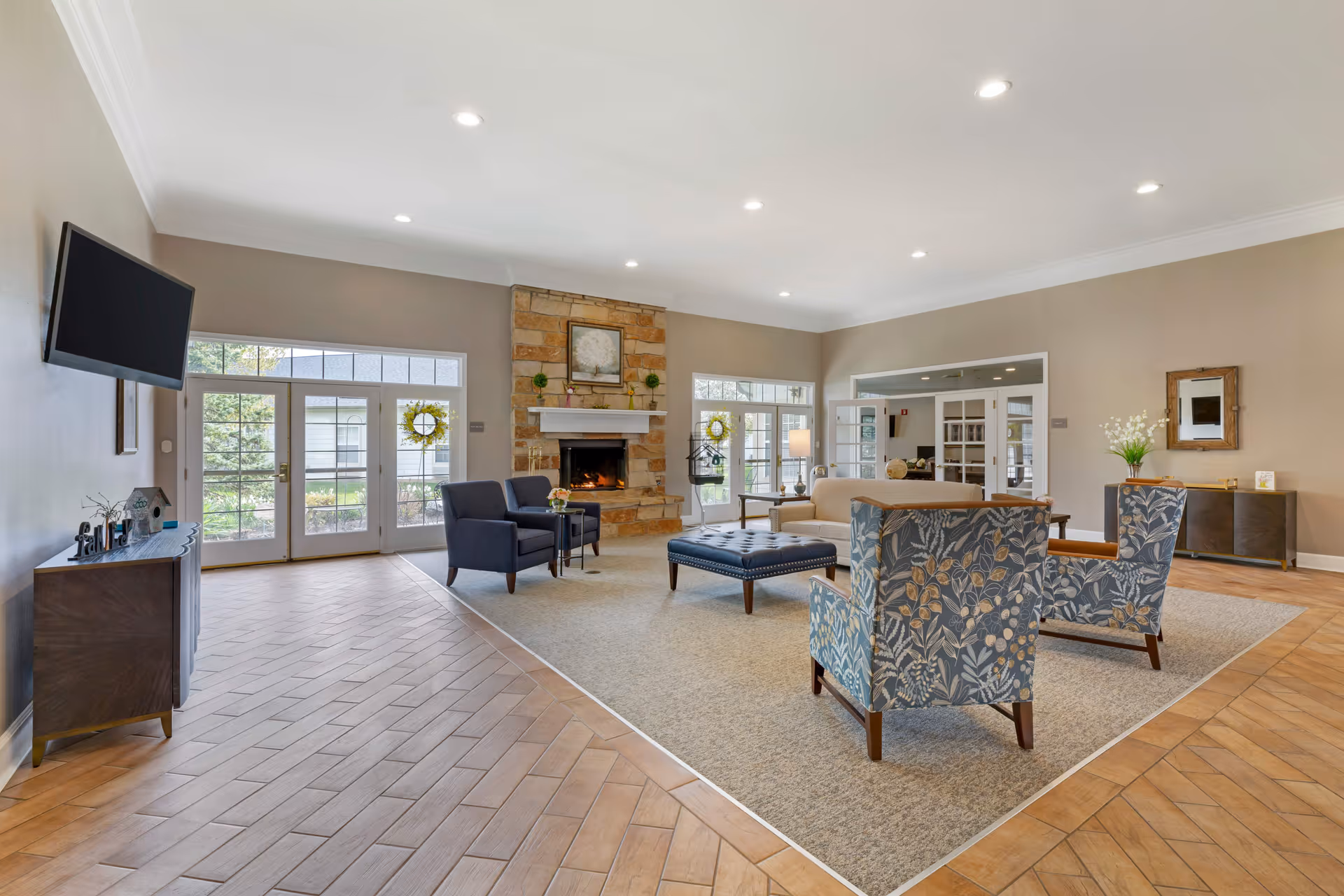 A spacious and well-lit senior living facility common area with a stone fireplace, multiple seating options including patterned armchairs, a beige sofa, and a blue ottoman. The room features large windows and glass doors letting in natural light, wood-patterned tile flooring, and neutral-colored walls with recessed ceiling lights.