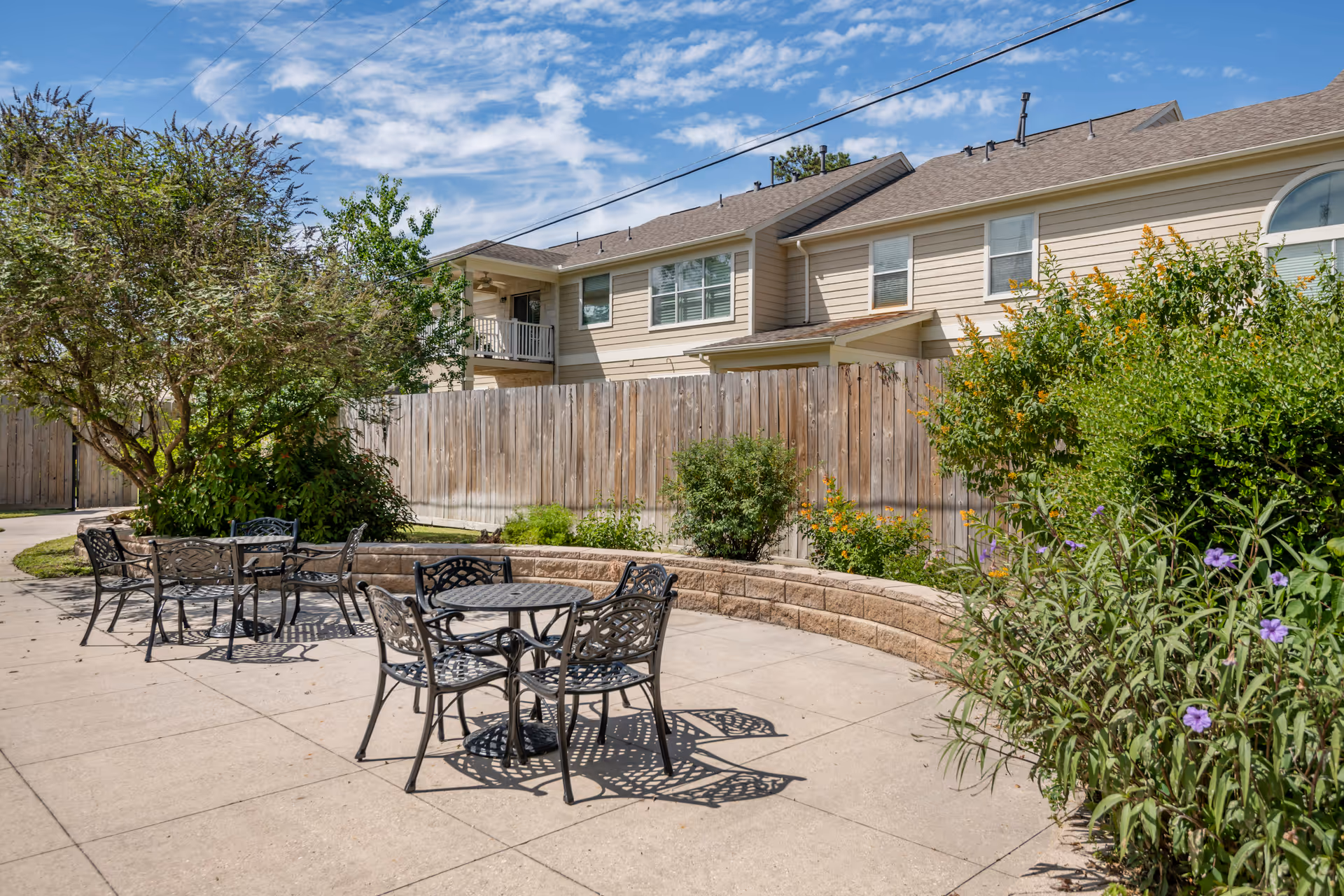 Outdoor patio area with metal tables and chairs on a concrete surface, surrounded by greenery and bushes. In the background, there is a wooden fence and a two-story beige residential building under a partly cloudy blue sky.