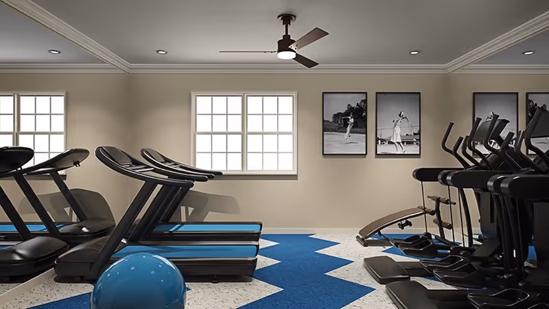 Interior view of a fitness room with several treadmills and elliptical machines arranged in rows. The room has beige walls, two windows letting in natural light, a ceiling fan, and three black and white framed photos of people playing tennis on the wall. A blue exercise ball is visible in the foreground on a blue and white patterned floor.