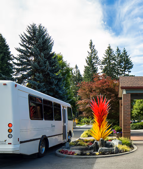 A white shuttle bus parked at the landscaped entrance with a tall red-and-yellow sculpture and trees in the background.