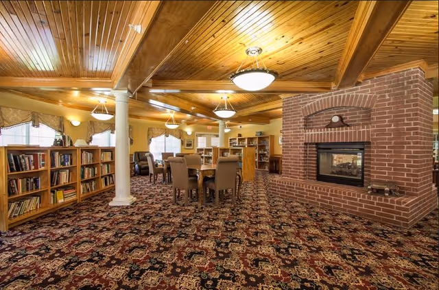 A cozy senior living community common area with a patterned carpet, wooden ceiling with beams, and warm lighting. The room features a brick fireplace, several bookshelves filled with books, and a table with chairs for seating. Large windows with curtains allow natural light to enter the space.