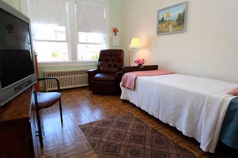 A senior living bedroom with a single bed, TV, chair, armchair, dresser, and window light.