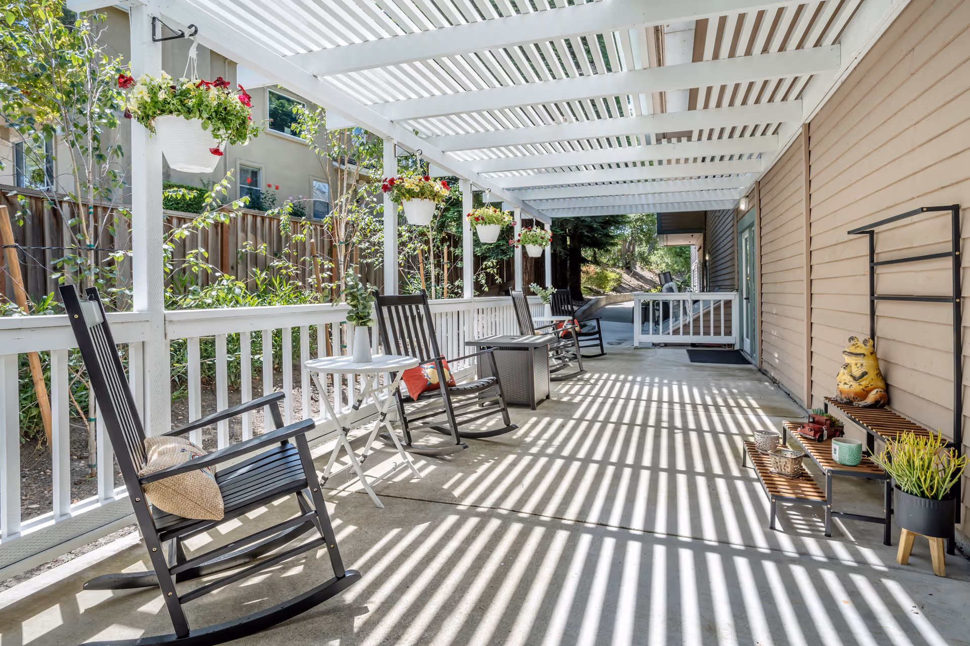 A covered outdoor patio area with white pergola casting striped shadows on the concrete floor. The patio features black rocking chairs with cushions, small white tables, hanging flower pots with red and white flowers, and a small wooden shelf with decorative items including a yellow frog statue and potted plants. The area is adjacent to a beige building with a wooden fence and greenery in the background.