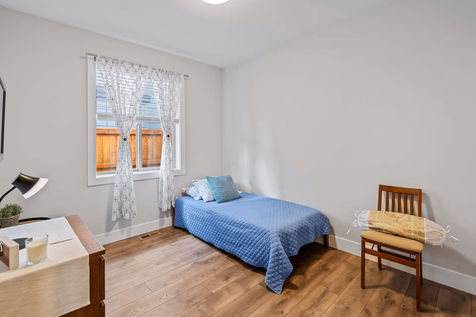 A simple bedroom with a single bed covered in a blue quilt and several pillows. There is a wooden chair with a decorative bolster pillow on it, a wooden dresser with a lamp, a small plant, and a candle. The room has light-colored walls, a window with sheer white curtains, and wooden flooring.