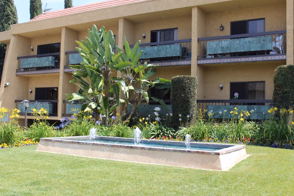 Exterior view of a two-story retirement hotel with balconies, landscaped gardens, and a rectangular fountain in the foreground.