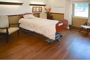 A single hospital-style bed with white bedding in a room with wooden flooring. The bed is positioned against a wall with a padded headboard and two chairs on either side. There is a window with white curtains letting in natural light, and a small table with a flower arrangement near the bed.