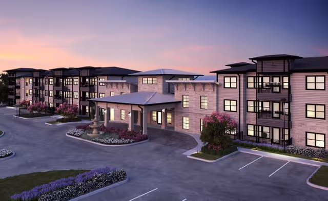 Exterior view of a modern senior living facility building at dusk with landscaped flower beds, a fountain, and a driveway in front.