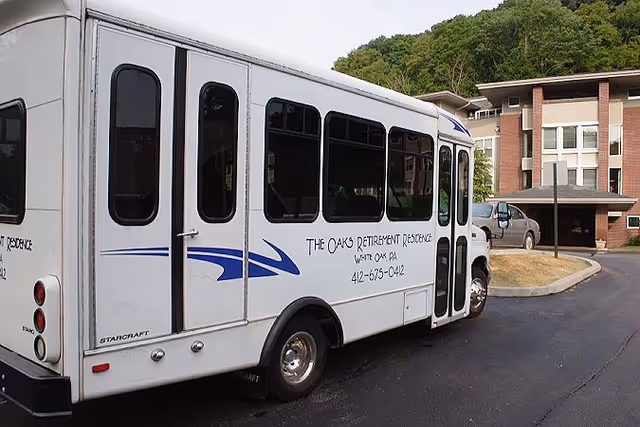 A white shuttle bus parked on a driveway in front of a multi-story brick and beige retirement residence building. The bus has blue decorative stripes and text on the side that reads 'The Oaks Retirement Residence, Wexford, PA, 412-675-0412'. There are trees and greenery in the background.