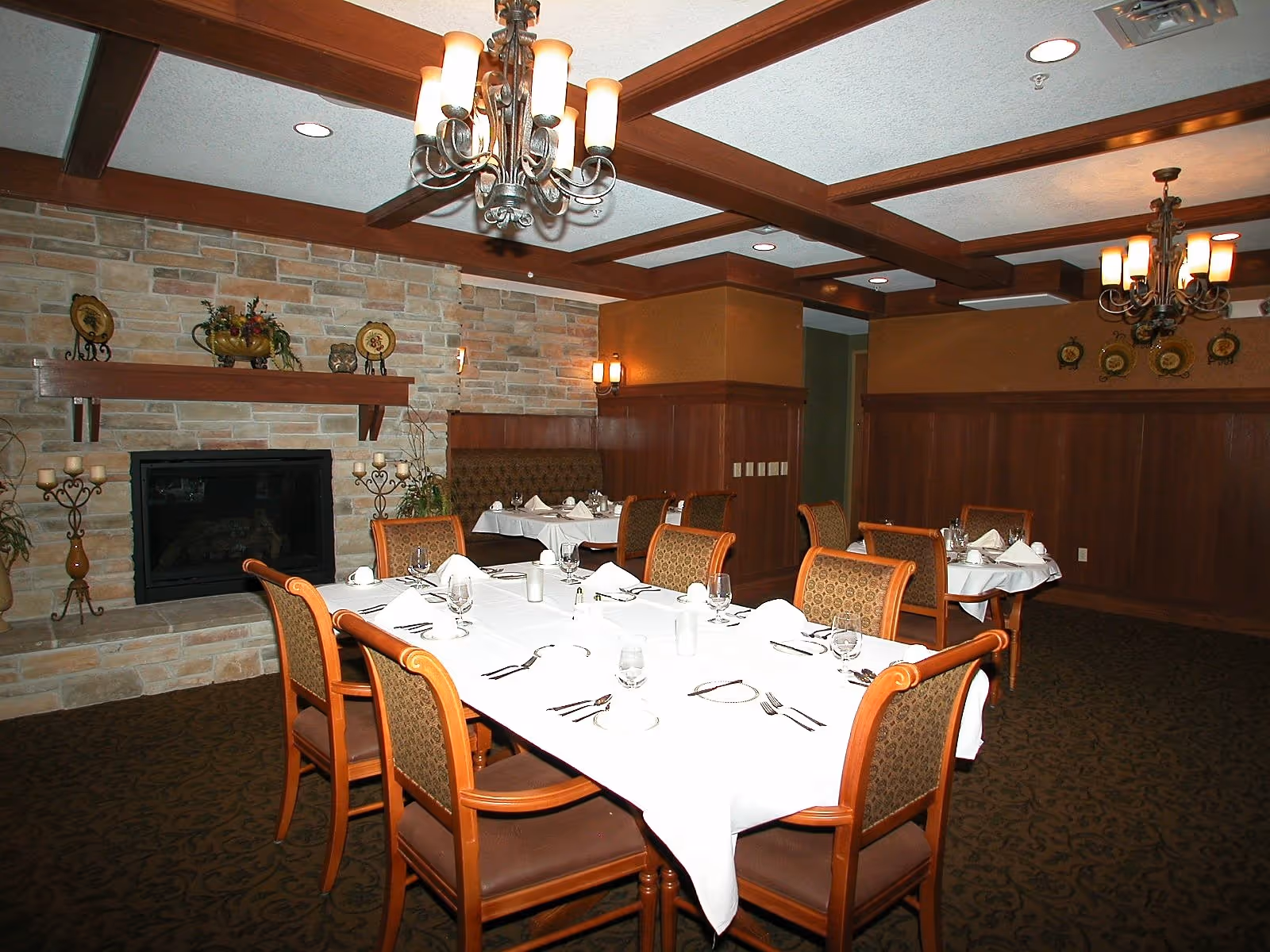 A dining room with a long table covered with a white tablecloth, set with glasses, plates, and silverware. The room features wooden chairs with patterned upholstery, a stone fireplace with decorative items on the mantel, wood-paneled walls, and ceiling beams with chandeliers providing warm lighting.
