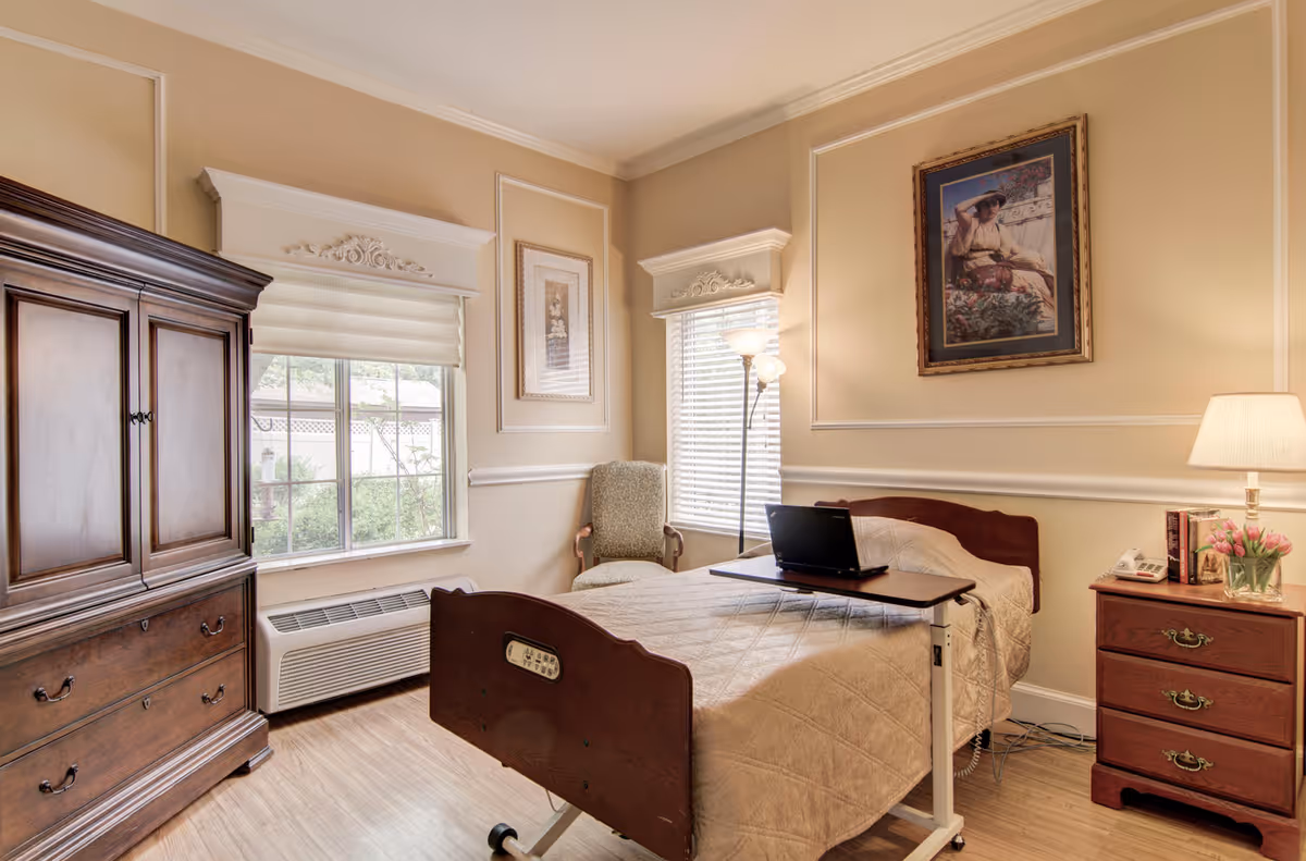 A cozy bedroom in a senior living facility featuring a single bed with a beige quilt, a wooden nightstand with a lamp, books, and a vase of pink flowers. There is a wooden armoire, a cushioned armchair near two windows with blinds, and a framed painting on the wall. A laptop is placed on a movable overbed table.