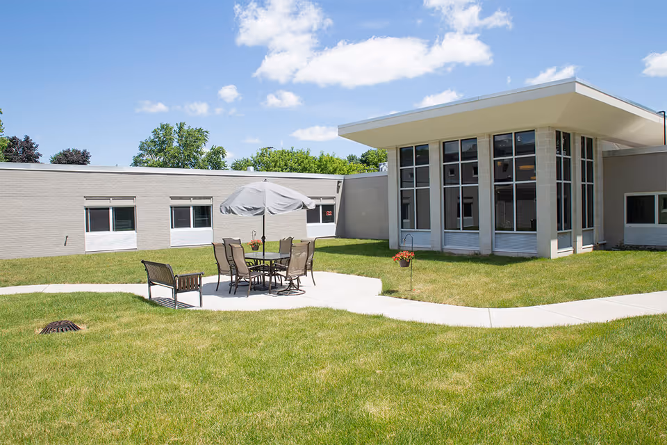 Outdoor courtyard area at Royal Oak House with green grass, a concrete pathway, and a patio set including a table with an umbrella and several chairs. The building has large windows and a flat roof under a blue sky with some clouds.