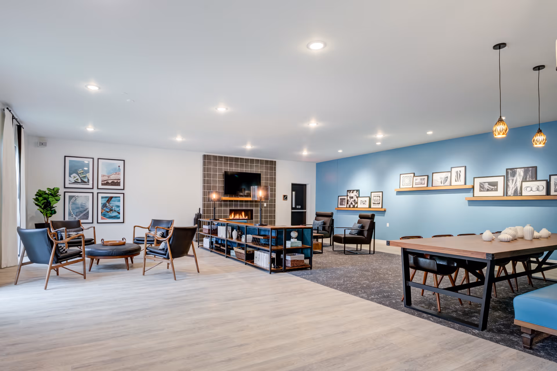 A spacious and modern living room area in a senior living facility named Mission Ridge, featuring a seating arrangement with four black chairs around a round coffee table on the left, a fireplace with a TV mounted above it in the center, and a long wooden table with chairs on the right. The walls are decorated with framed pictures and shelves, and the room has a light wood floor and a blue accent wall.
