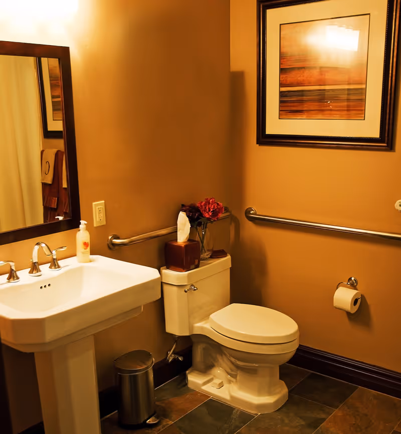 A bathroom with beige walls featuring a white pedestal sink with a soap dispenser, a white toilet with a tissue box and a vase with red flowers on top, a metal trash can, a wall-mounted handrail, a toilet paper holder with a roll of toilet paper, a framed abstract artwork on the wall, and a mirror reflecting part of the room.