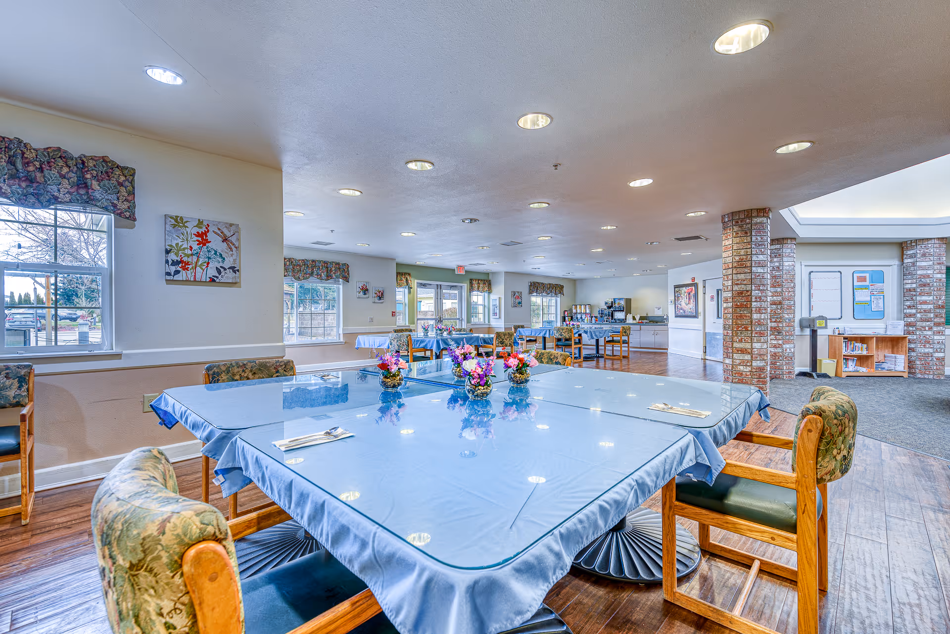 A spacious dining room in an assisted living facility with tables covered in blue tablecloths and glass tops, each decorated with small floral centerpieces. The room has wooden chairs with floral upholstery, large windows with floral valances, and bright ceiling lights. In the background, there is a beverage station and a bulletin board near brick columns.