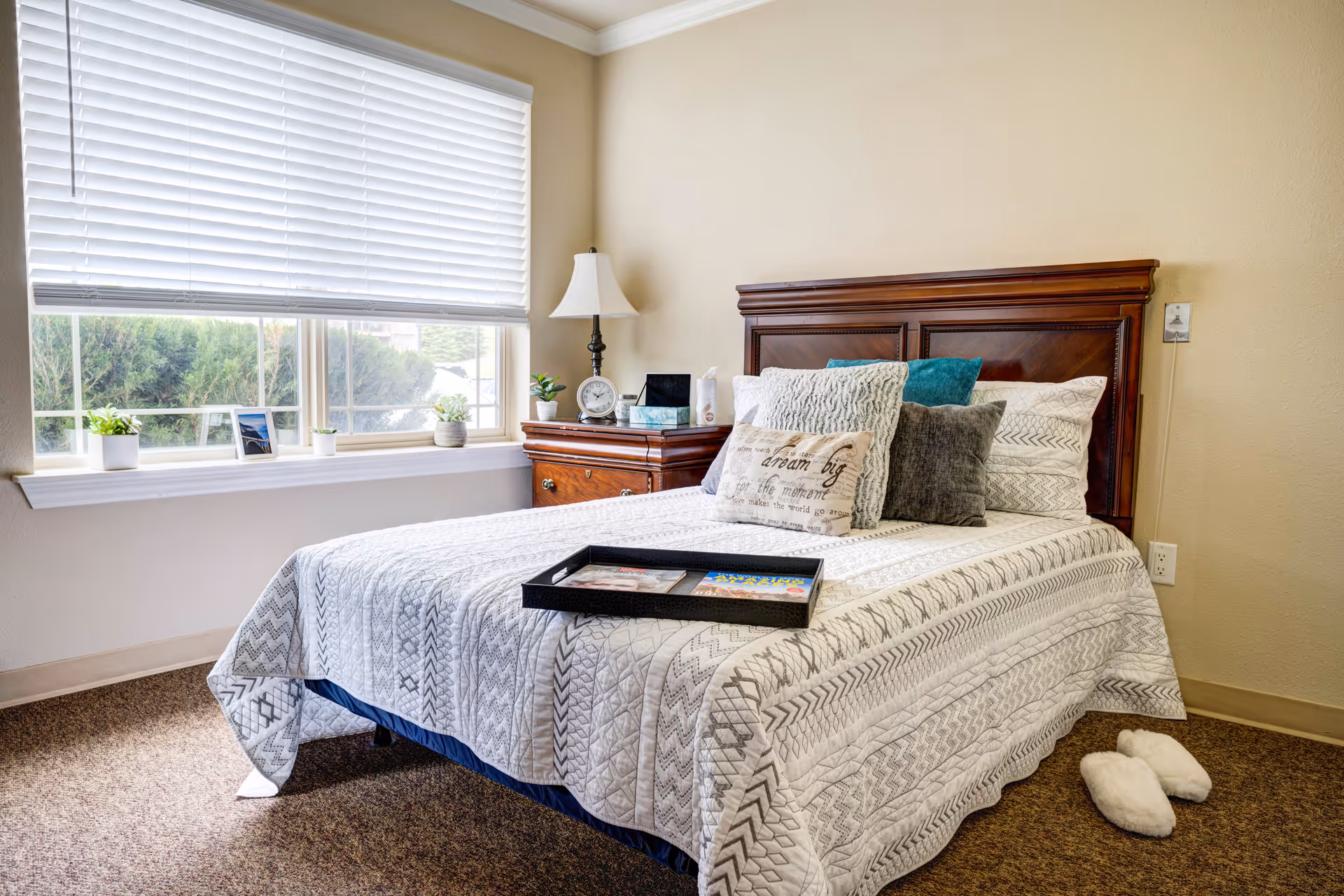 A cozy bedroom with a neatly made bed featuring a white and gray patterned quilt and multiple decorative pillows. A wooden headboard and matching nightstand with a lamp, clock, and small plants are beside the bed. A tray with magazines is placed on the bed. A large window with white blinds lets in natural light, and a pair of white slippers is on the carpeted floor next to the bed.