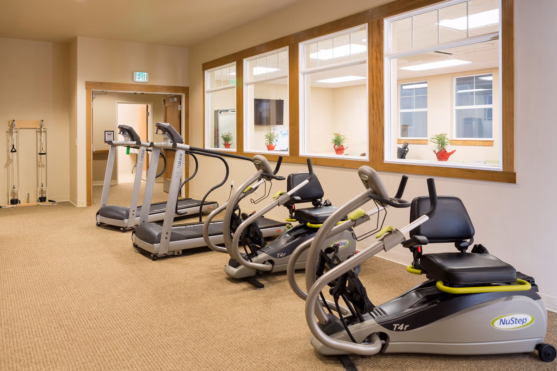 A fitness room with two treadmills and two recumbent exercise bikes on a carpeted floor. The room has beige walls and large windows looking into an adjacent room with potted plants on the windowsills. There is an exit door at the back and some exercise equipment mounted on the wall.