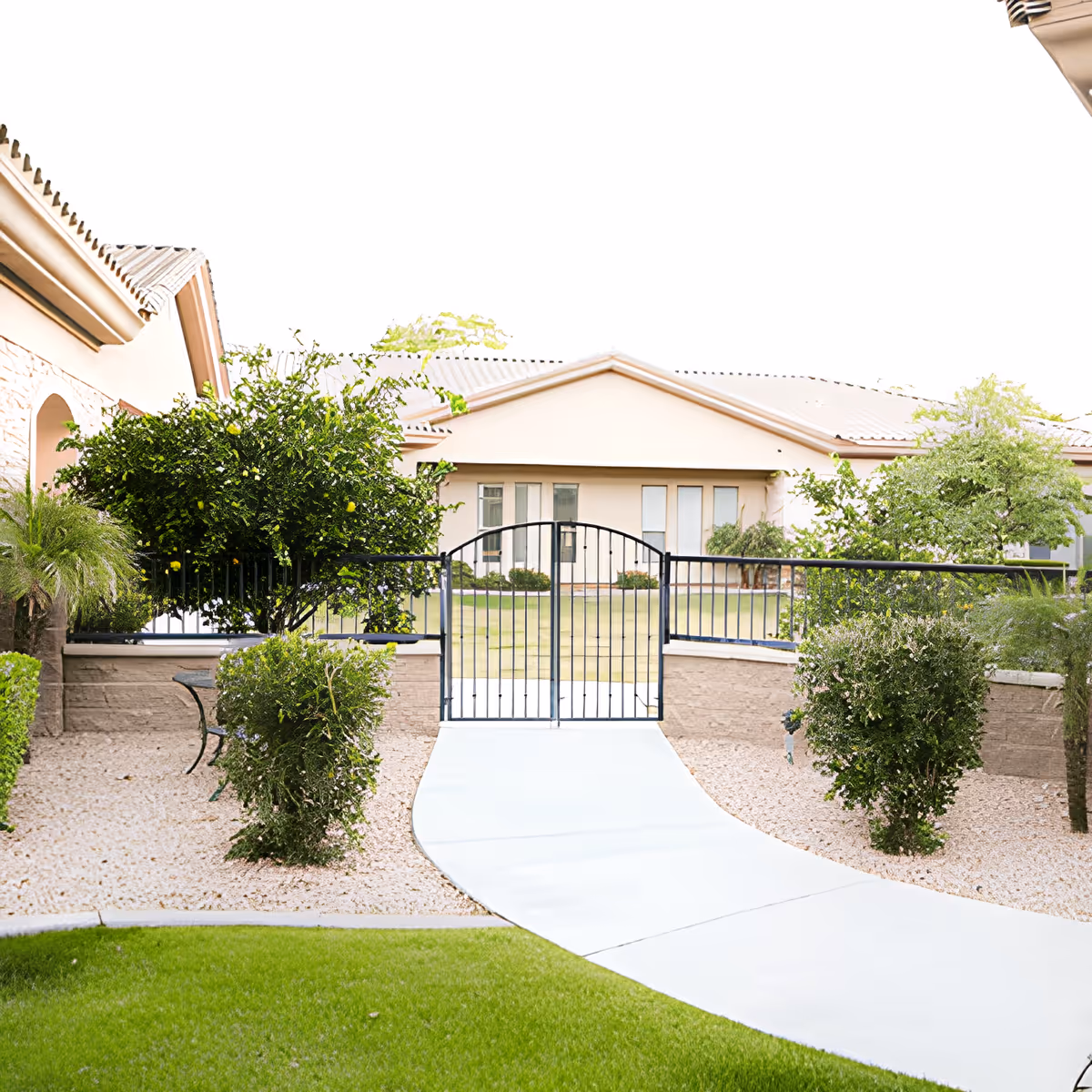 A curved paved walkway leads through gravel landscaping and shrubs to a black metal gate opening to a single-story building with a tiled roof.