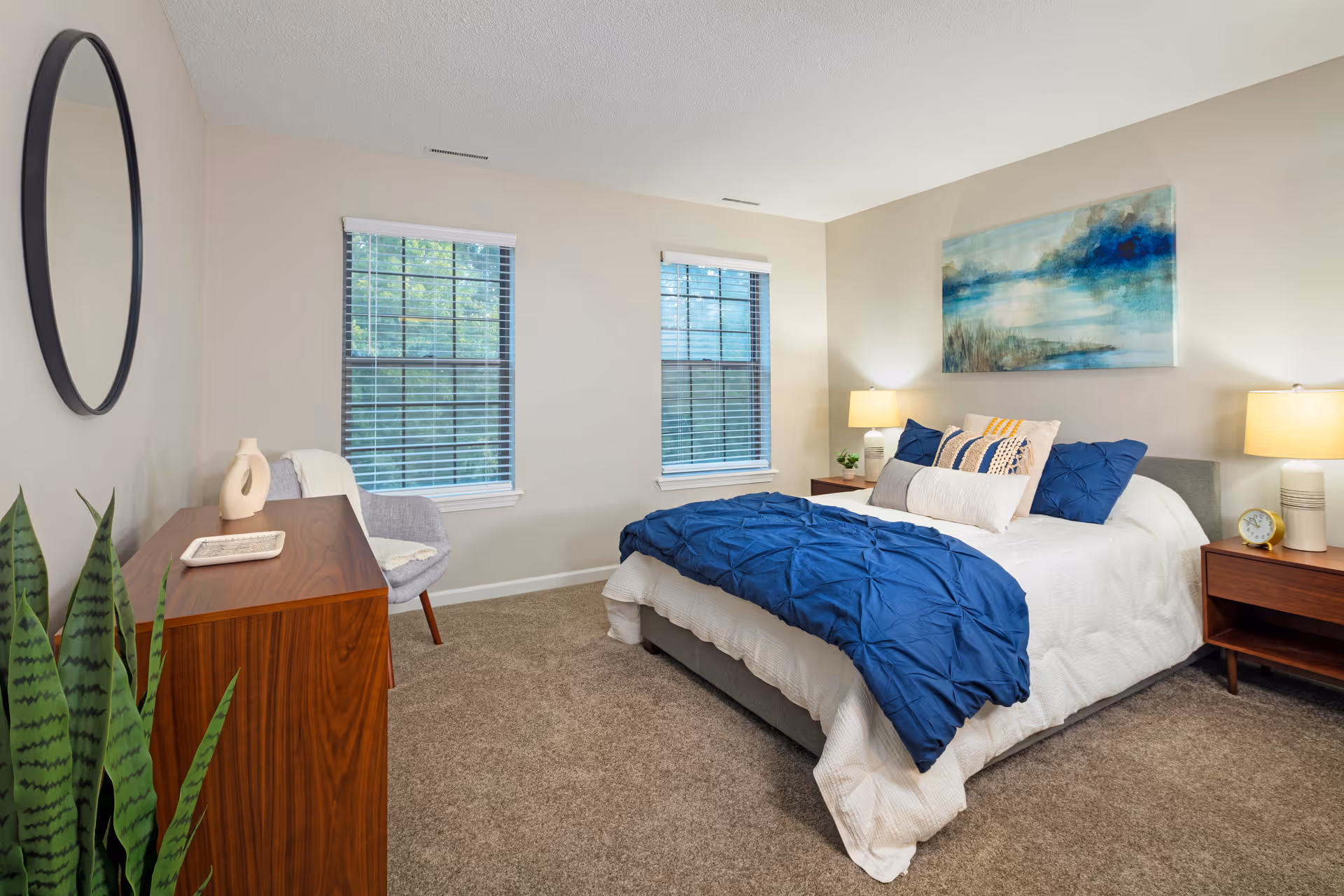A neatly arranged bedroom with a bed covered in white and blue bedding, two wooden nightstands with lamps on either side, a wooden dresser with decorative items, a gray chair, two windows with blinds, a round mirror on the wall, a large painting above the bed, and a green plant in the corner.