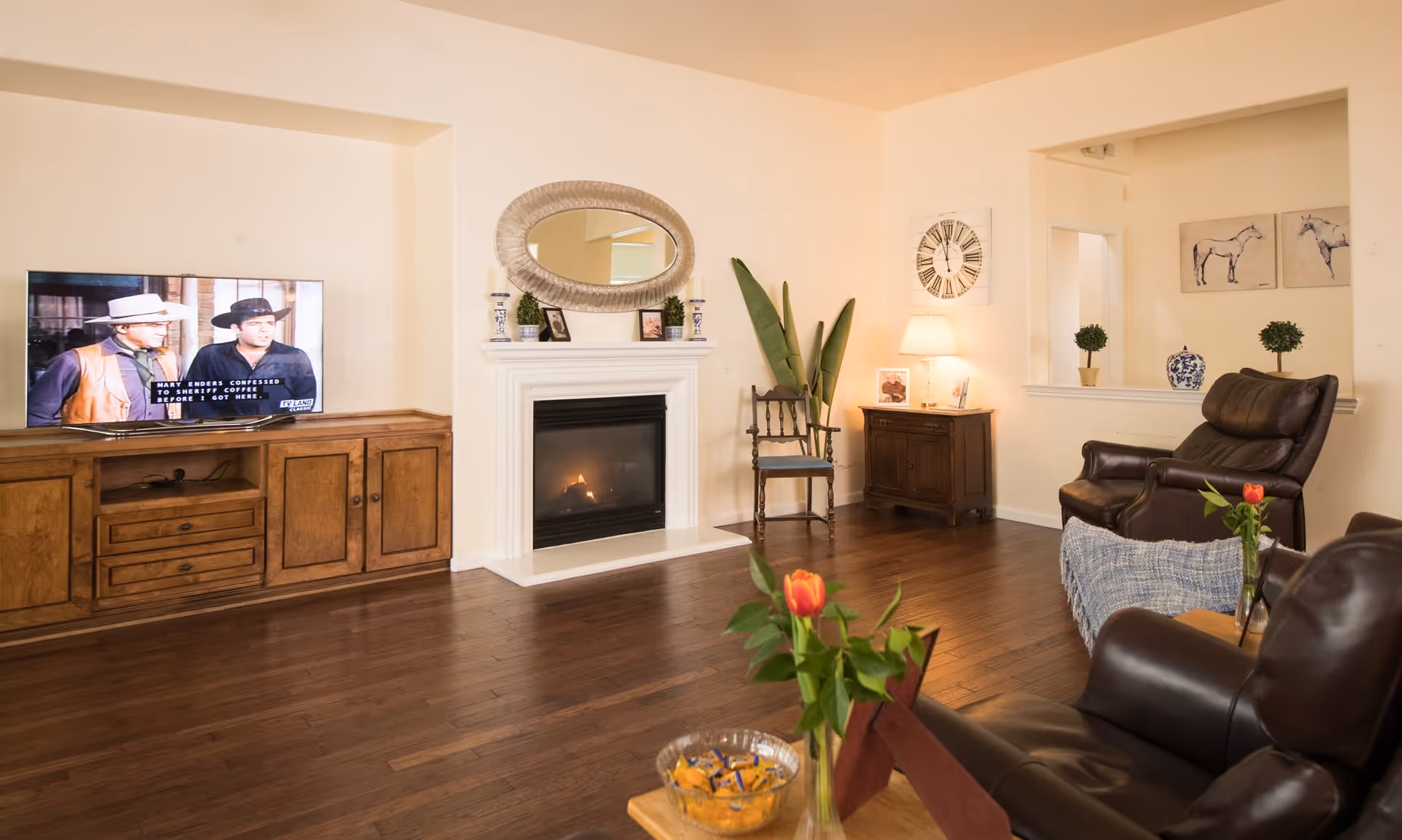 A cozy senior living room with wooden flooring, a fireplace with a decorative mirror above it, a flat-screen TV on a wooden cabinet showing a western movie, two leather armchairs, a wooden chair, a small wooden cabinet with a lamp, and decorative plants and artwork on the walls.