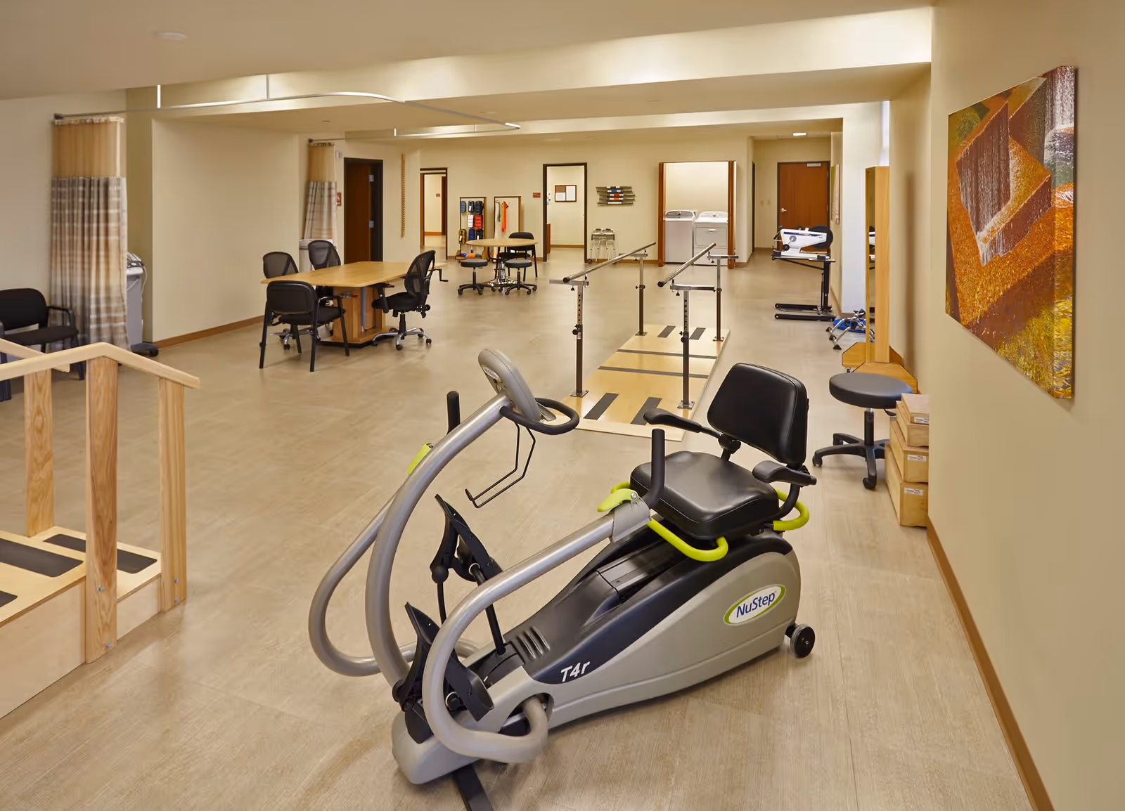 A spacious rehabilitation or physical therapy room with exercise equipment including a NuStep recumbent cross trainer, parallel bars for walking exercises, tables with chairs, and various therapy tools. The room has light-colored walls and flooring, with a colorful abstract painting on one wall.