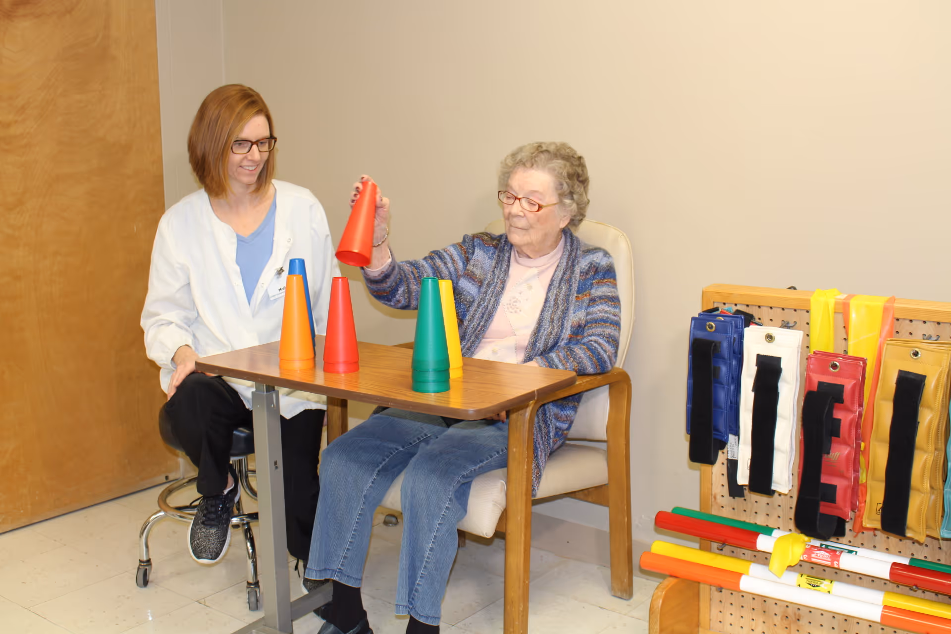 An elderly woman sitting in a chair at a table stacking colorful plastic cones while a caregiver or therapist watches and smiles. Various therapy or exercise equipment is visible on a pegboard nearby.