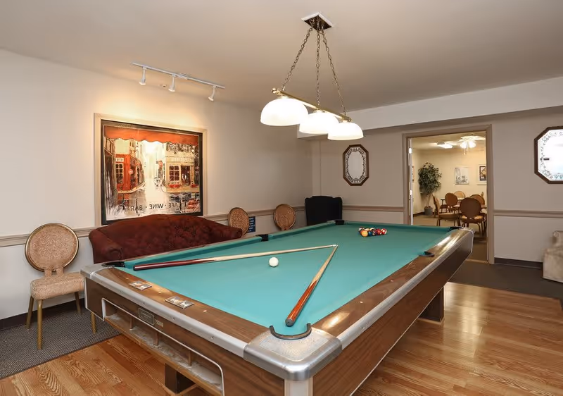 A recreational room with a pool table in the center, two pool cues resting on the table, a red couch against the wall, two chairs, a framed painting above the couch, and a hanging light fixture above the pool table. An adjacent room with a table and chairs is visible through an open doorway.