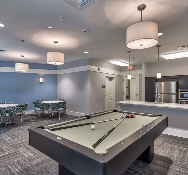 Interior view of a senior living facility common area featuring a pool table with cues and balls set up, a kitchen area with stainless steel refrigerator and microwave, and a dining area with round tables and patterned chairs under modern pendant lights.