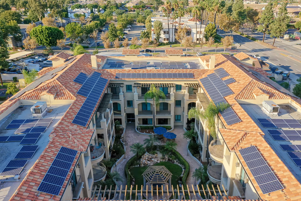 Aerial view of a U-shaped senior living building with terracotta roofs covered in solar panels surrounding a landscaped central courtyard with palm trees and a gazebo.