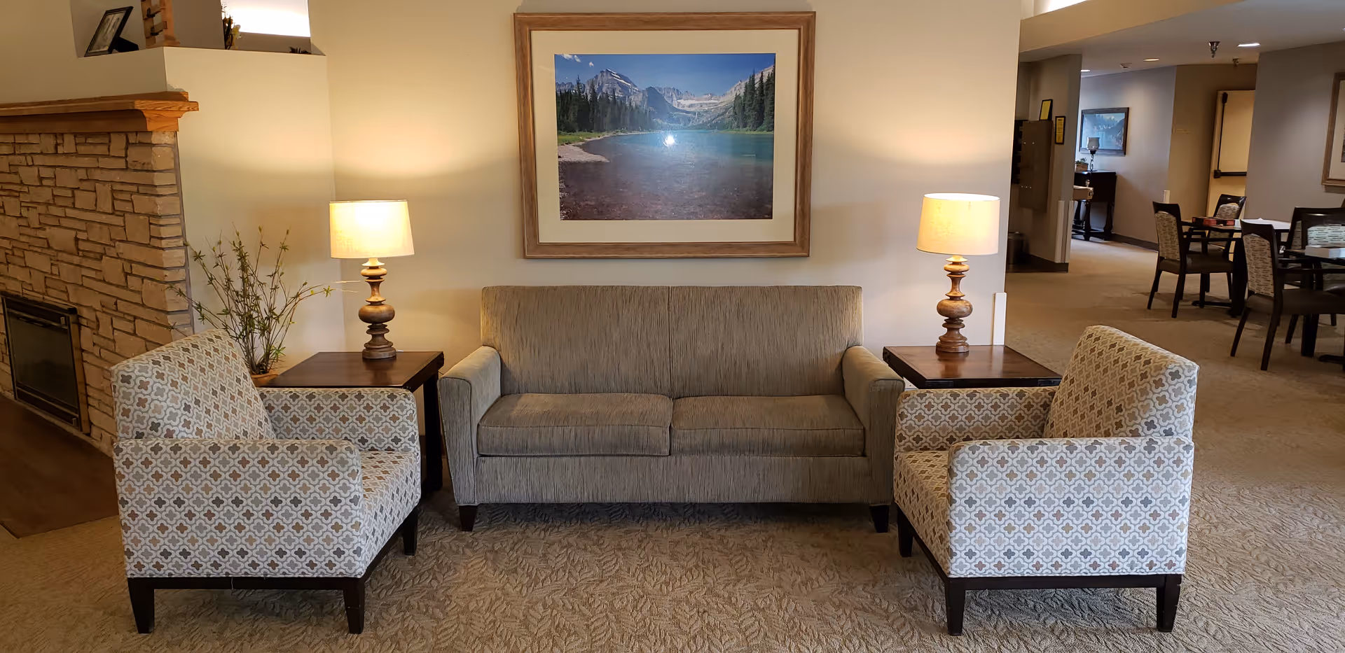 A cozy seating area in a senior living facility featuring a beige couch flanked by two patterned armchairs. Each side of the couch has a wooden side table with a lamp. Behind the couch is a large framed picture of a mountain lake landscape. To the left, there is a stone fireplace with a plant next to it. In the background, there is a dining area with tables and chairs.