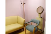 A small room with a yellow couch, a blue armchair, a wooden coat rack, and a vintage hair dryer machine against a light purple wall.