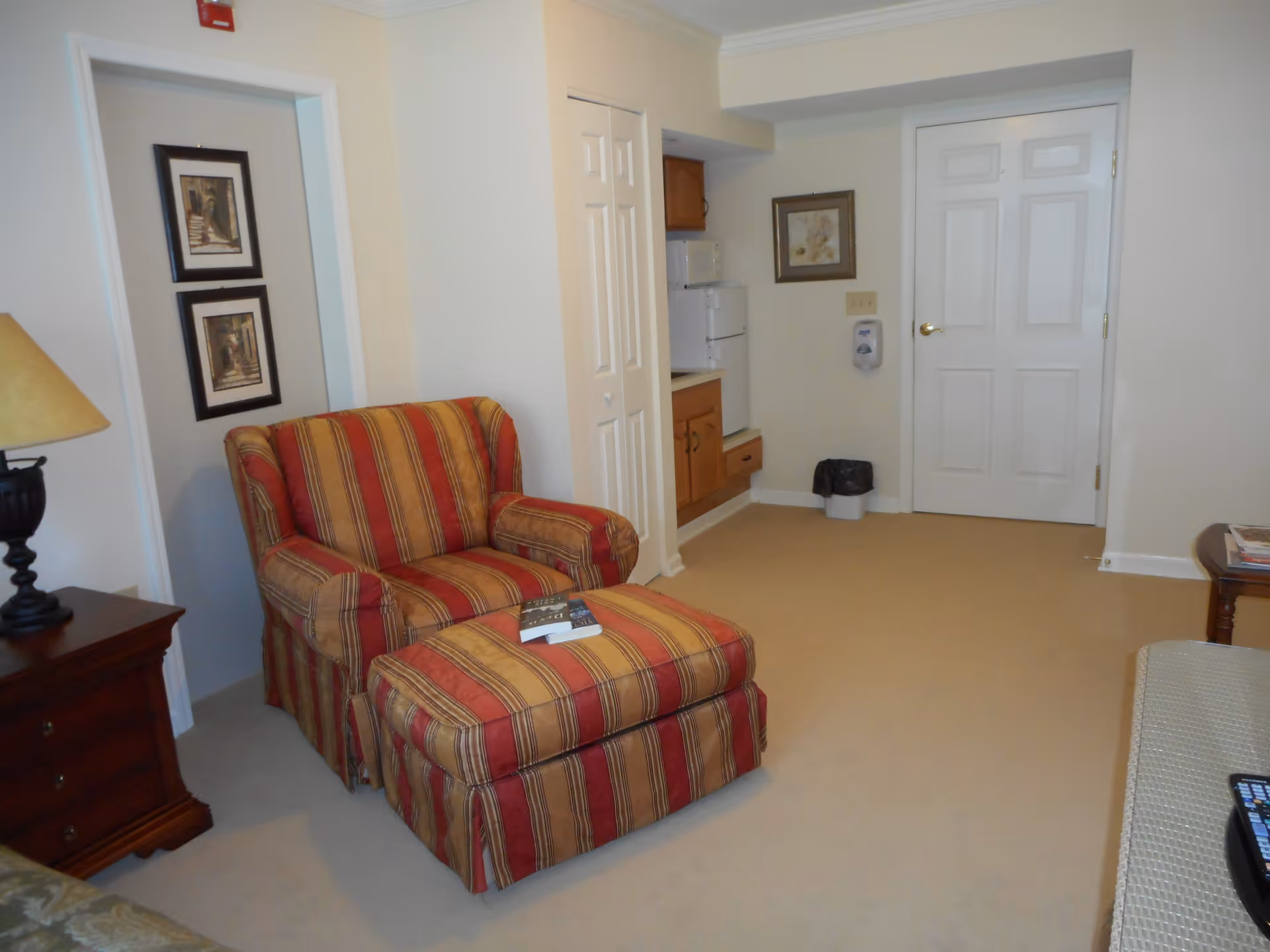 Interior view of a senior living facility room at Feridean Commons featuring a red and yellow striped armchair with matching ottoman, a wooden side table with a lamp, framed artwork on the wall, a small kitchenette area with a refrigerator and microwave, and a closed white door.