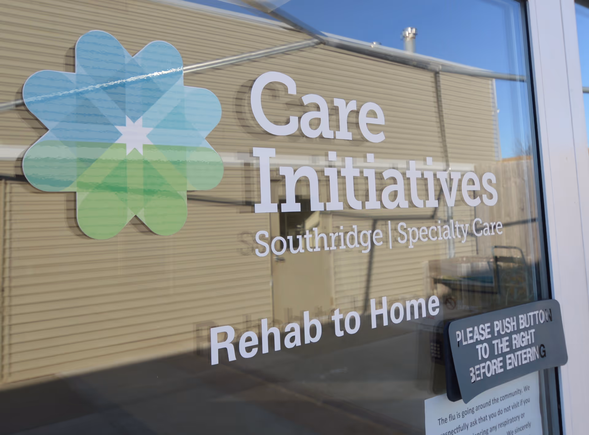 Glass door with signage for Care Initiatives Southridge Specialty Care Rehab to Home, with a colorful flower-like logo and a sign instructing to push the button to the right before entering.