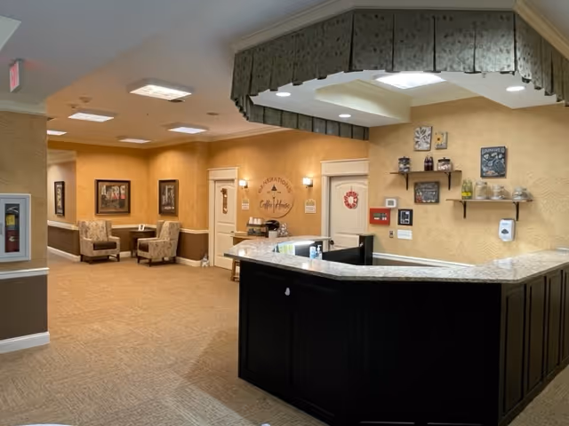 Interior view of a senior living facility reception area with a dark wood front desk and a beige countertop. Behind the desk, there are shelves with decorative items and a sign that reads 'Generations Coffee House.' The walls are painted in warm beige tones with framed artwork and two upholstered chairs with a small table in a seating area to the left.