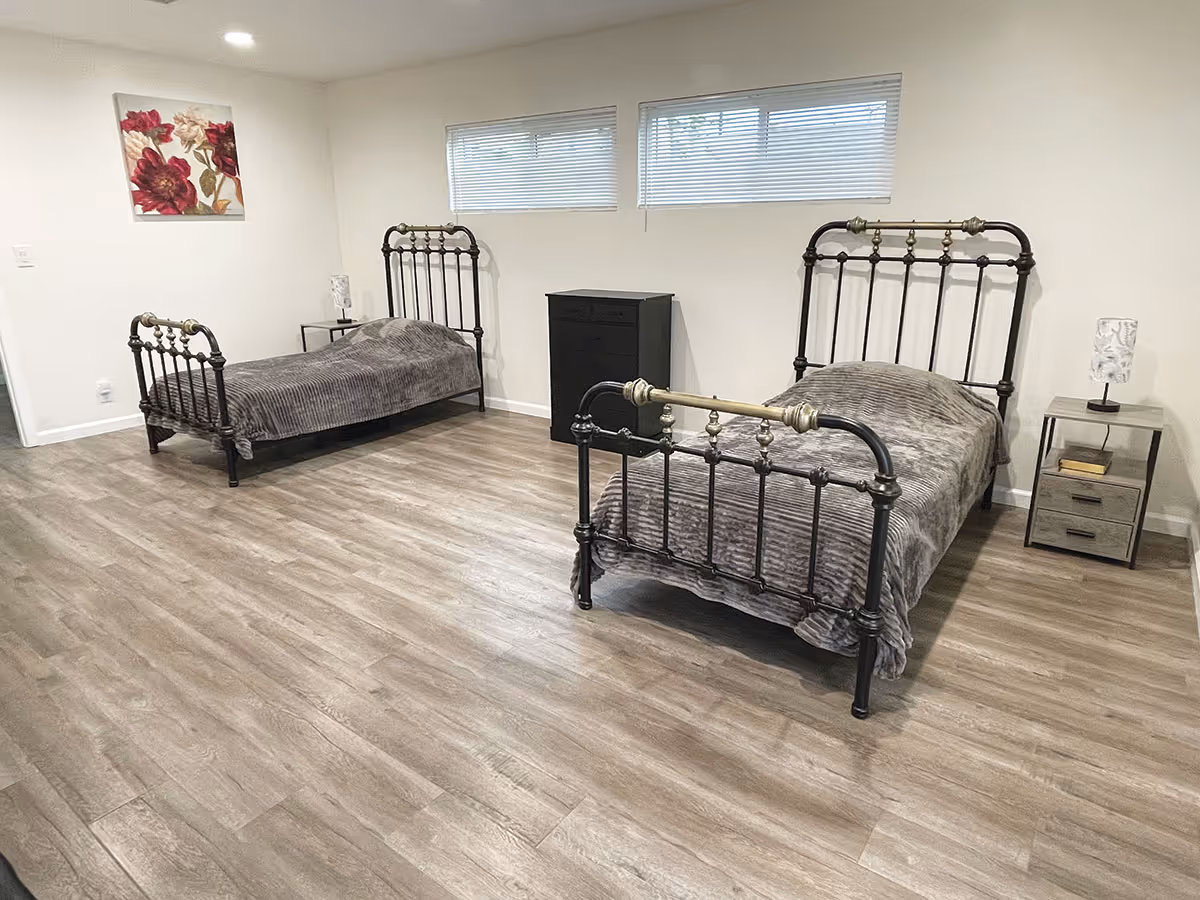 A bedroom with two single metal frame beds covered with gray blankets. Each bed has a nightstand with a lamp, and there is a black dresser between the beds. The room has light-colored walls, wood flooring, two small windows with blinds, and a floral painting on one wall.