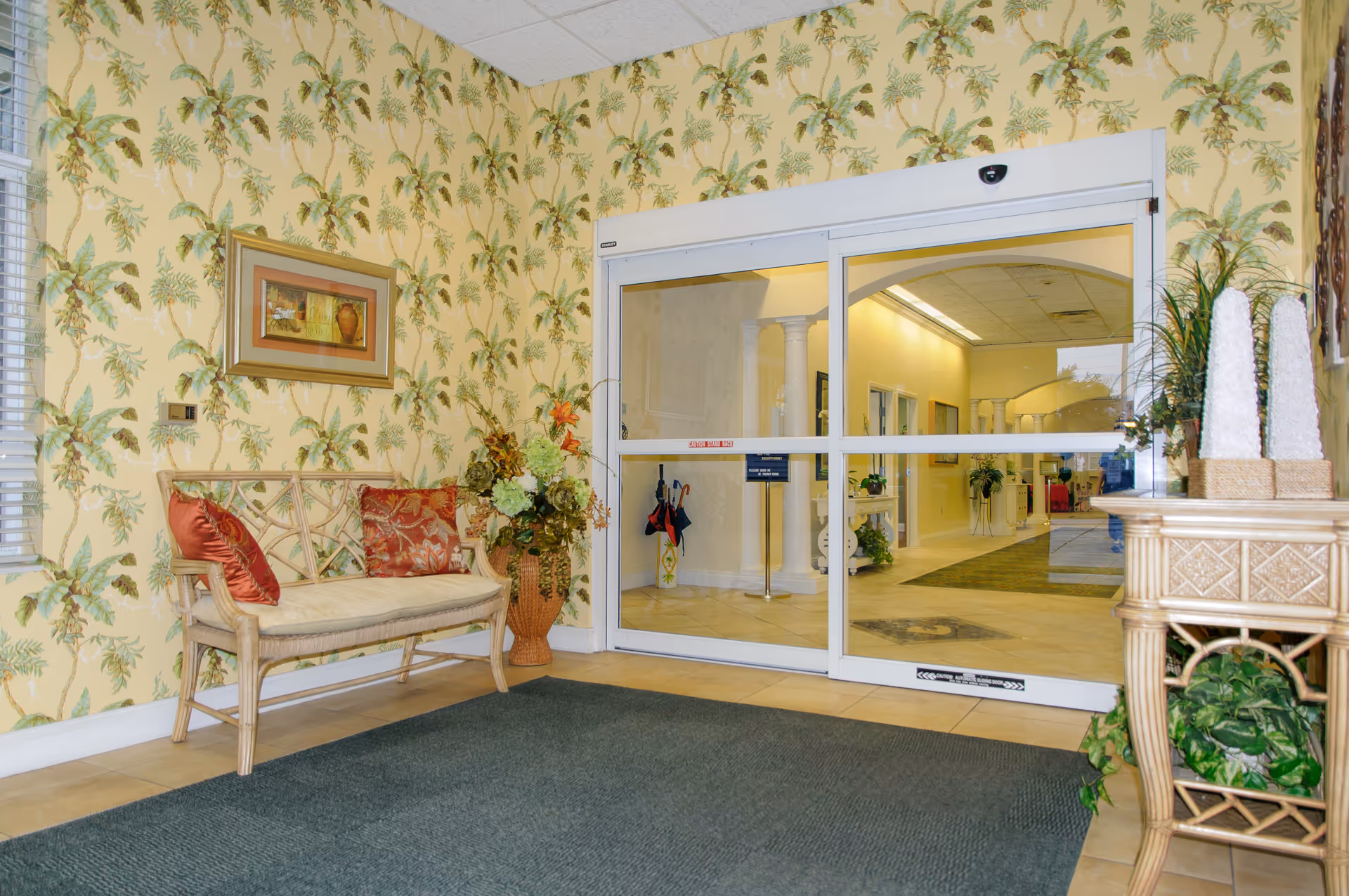 Entrance area of a senior living facility with a glass automatic sliding door. There is a wicker bench with red and patterned cushions on the left side, a large floral arrangement in a vase next to it, and a wicker table with decorative items and plants on the right. The walls have yellow wallpaper with a green leafy pattern, and a framed picture hangs above the bench. The floor has a dark mat near the door and light-colored tiles.