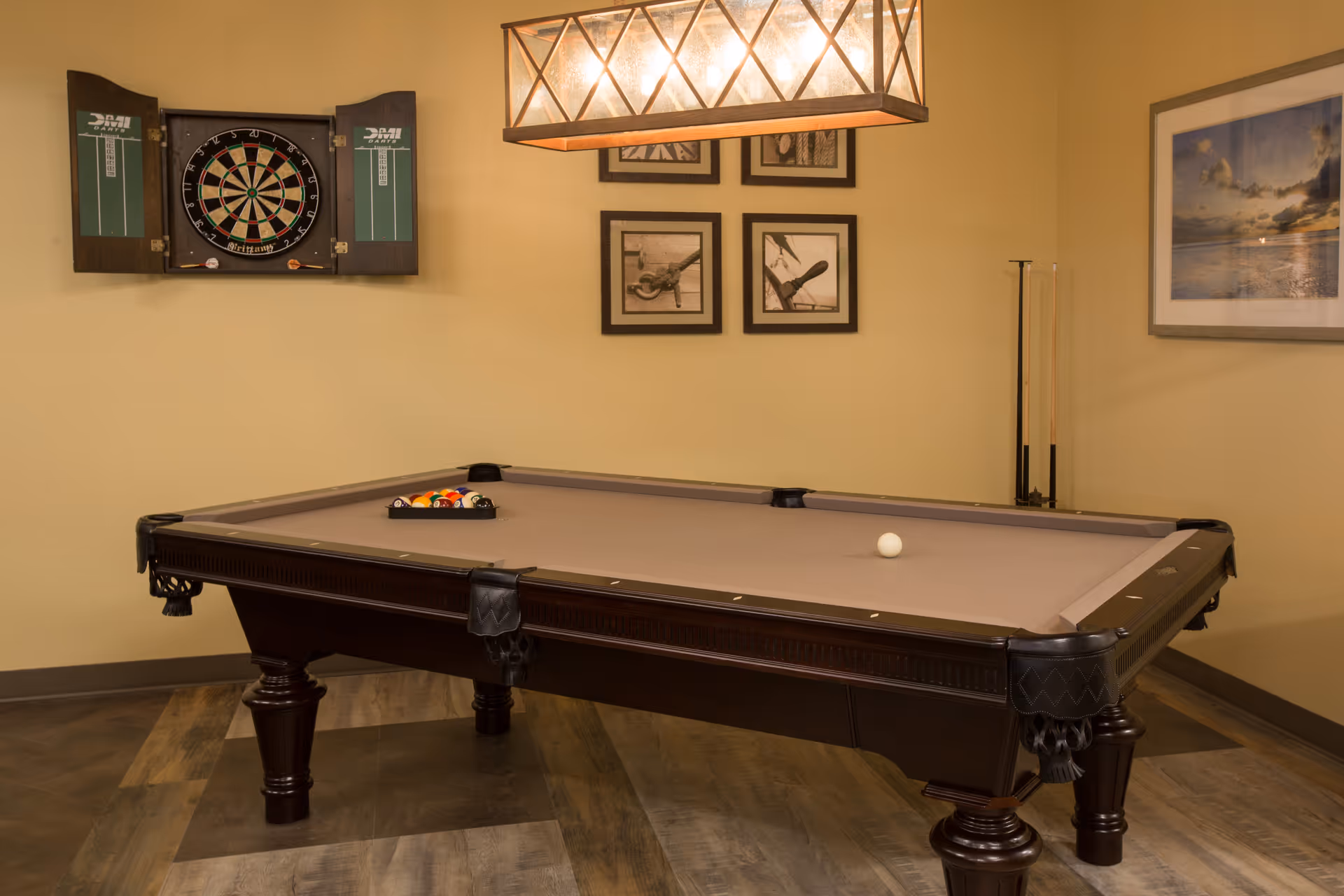 A game room with a pool table, a wall-mounted dartboard, pool cues, and framed artwork under a hanging light.