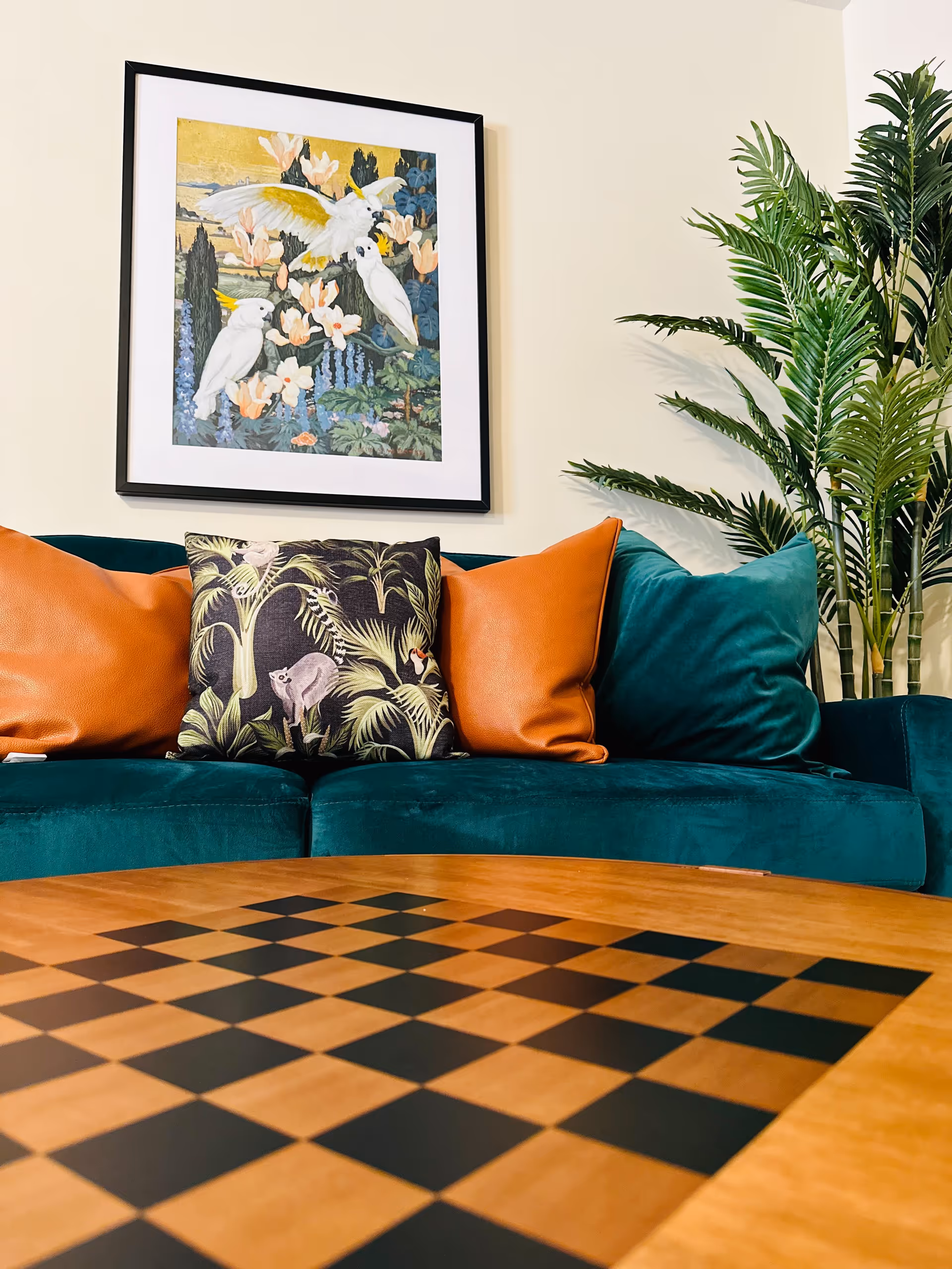 Teal velvet sofa with colorful throw pillows beneath framed bird artwork, a potted plant to the right, and a checkerboard table in the foreground.